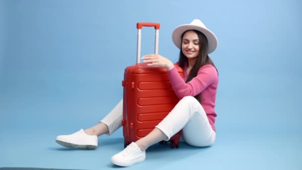 Šťastná mladá turistka sedící u kufru, objímající ho a cestující na dovolené izolované na modrém pozadí - Záběry, video