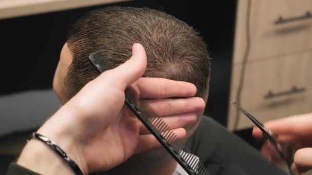 El estilista corta el cabello del cliente con tijeras afiladas que sostienen el peine en primer plano de la peluquería. Maestro hace corte de pelo clásico a caballero en salón de belleza - Metraje, vídeo