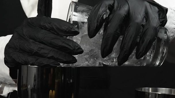 Makrographie, erleben Sie die geschickten Hände eines Barkeepers, wie sie gekonnt einen Cocktail mit schwarzem Hintergrund mixen. Nahaufnahme erfassen das Pour Ice in Shaker und bereiten für die Herstellung von Cocktail. Komestibel. - Foto, Bild