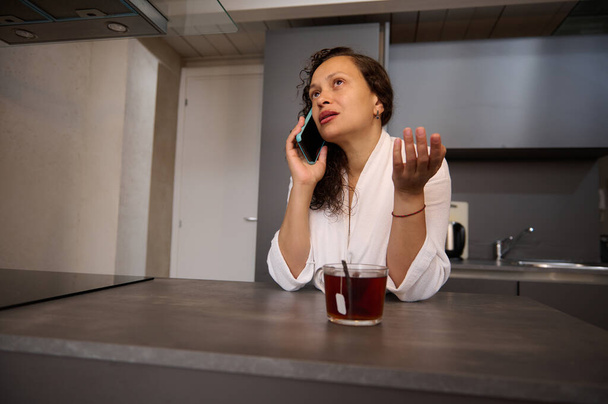 Hermosa mujer joven en albornoz blanco, hablando por teléfono móvil sobre una taza de té caliente en la mañana - Foto, Imagen