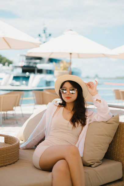 Die schöne junge Asiatin sitzt im Café am Hafen und hält ihren Strohhut mit verschwommener Jacht und Meer im Hintergrund. Sie trägt ein schmales cremefarbenes Kleid, rosa und reflektierende Sonnenbrille, Strohhut und Halskette. - Foto, Bild