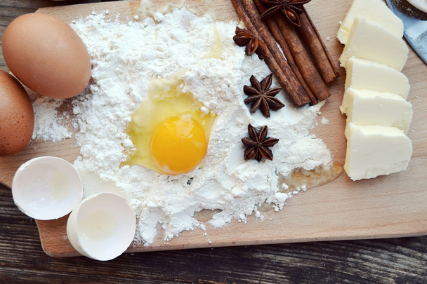 White flour with eggs, butter, cinnamon sticks, spicy anise stars and wooden spoon on a cooking board - Photo, Image