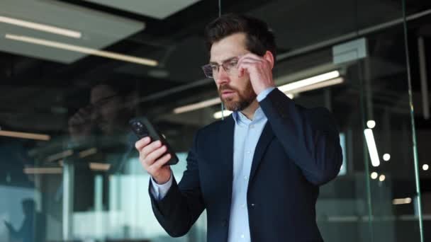 Empresário caucasiano surpreso removendo óculos e olhando para a exibição de smartphone pessoal com expressões faciais de choque. Punchy masculino vestido em trajes formais escuros em pé no ambiente de escritório. - Filmagem, Vídeo