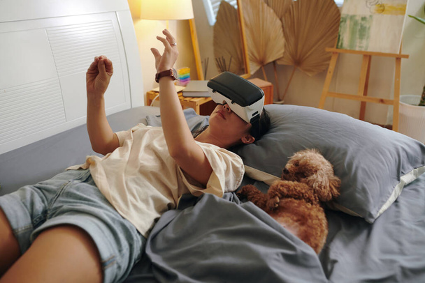 Mujer joven acostada en la cama junto a un perrito y jugando en gafas vr - Foto, imagen