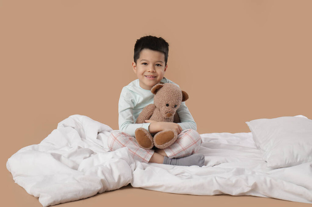 słodkie mały Azji chłopiec w piżama z zabawka niedźwiedź poduszka i koc siedzi na beżowy tło - Zdjęcie, obraz