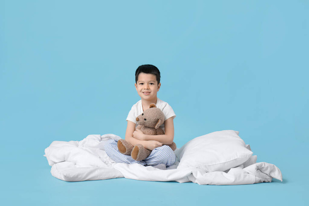 Pijamalı, oyuncak ayı, yastık ve battaniyeli şirin Asyalı çocuk mavi arka planda oturuyor. - Fotoğraf, Görsel