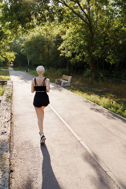 Vue arrière d'une jeune femme en tenue de sport faisant du jogging seule sur un sentier du parc avec des arbres et de la lumière du soleil, exprimant la santé et la forme physique. - Photo, image