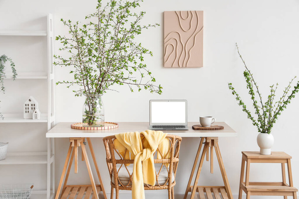 Стильне робоче місце, крісло, ноутбук і ваза з квітучими гілками у світлій вітальні - Фото, зображення