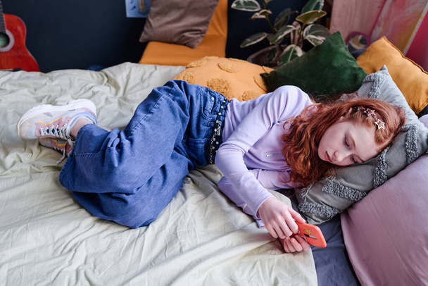 Vista de ángulo alto de la chica adolescente caucásica acostada en la cama en su dormitorio sosteniendo el teléfono inteligente esperando un mensaje o una llamada telefónica - Foto, imagen