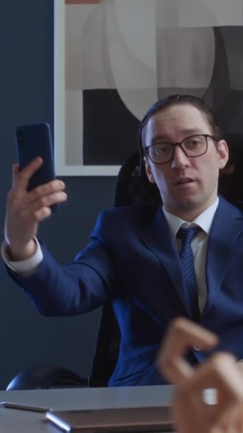Середня вертикаль нарцисичного чоловічого корпоративного боса в костюмі, окуляри, сидячи за столом, попереджаючи, маючи відеодзвінок на смартфон, захоплюючись собою, знущаючись, глузуючи з нервового колеги - Кадри, відео