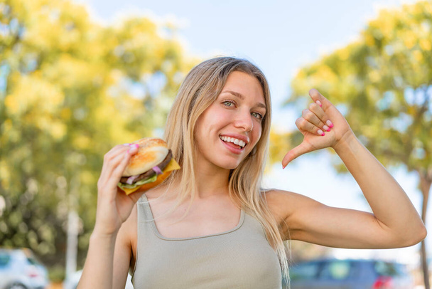 Joven rubia sosteniendo una hamburguesa al aire libre orgullosa y satisfecha - Foto, imagen