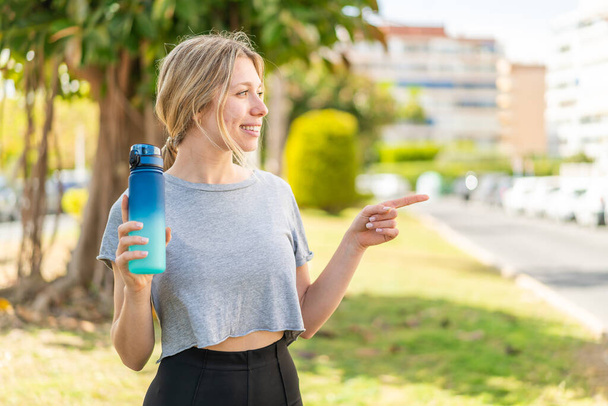 Молодая блондинка спортивная женщина с бутылкой воды на открытом воздухе, указывая в сторону представить продукт - Фото, изображение