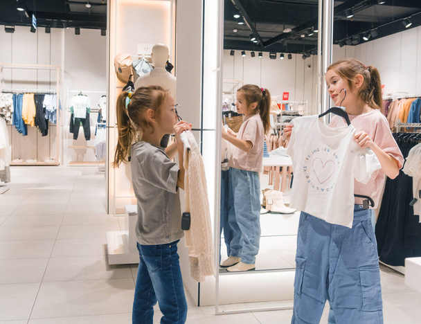 Deux filles, probablement des adolescents, parcourent des étagères de vêtements dans un magasin occupé rempli de divers vêtements. - Photo, image