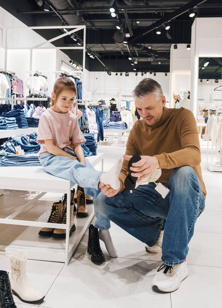 Gülümseyen bir kız, babası ona bir dükkanda yeni ayakkabılarla yardım ederken oturur.. - Fotoğraf, Görsel