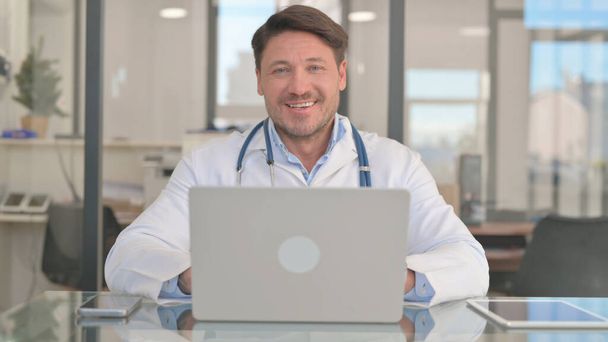 Arzt lächelt während der Arbeit am Laptop in die Kamera - Foto, Bild