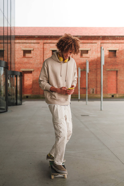 Jonge man met krullend haar skateboarden en met behulp van mobiele telefoon. Hoge kwaliteit foto - Foto, afbeelding