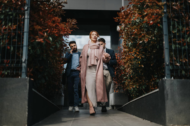 Elegante mujer de negocios avanza con confianza fuera de un edificio moderno, con colegas masculinos siguiendo. - Foto, imagen