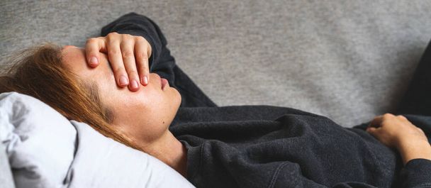 Η κουρασμένη κουρασμένη γυναίκα ξαπλώνει στον καναπέ και καλύπτει το πρόσωπό της με το χέρι της.. - Φωτογραφία, εικόνα