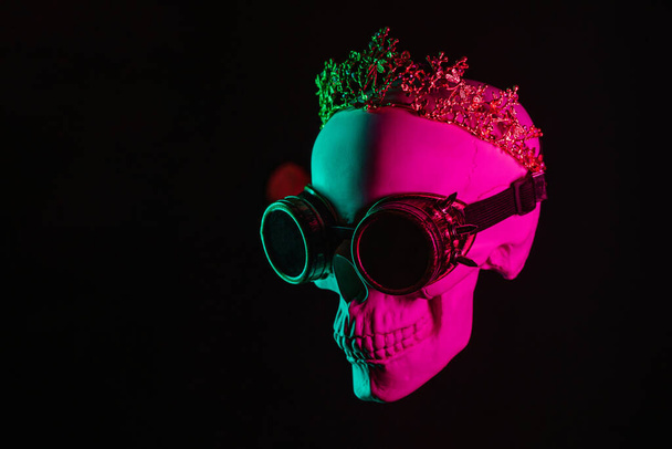 human skull in steampunk glasses and crown with pink green light on black background - Photo, Image
