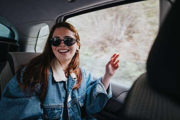 Jovem alegre usando óculos de sol, confortavelmente sentada em um carro, sentando o vento e expressando felicidade durante uma viagem de carro. - Foto, Imagem