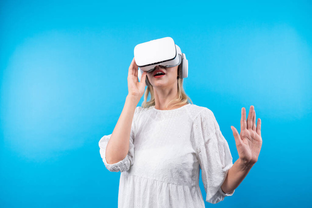 Mutlu kadın VR gözlüklerine bakıp mavi arka planda dururken şaşırdı ya da heyecanlandı. Beyaz pijamalı beyaz bir kız metaevrene bağlanmak için görsel gerçeklik gözlüğü kullanıyor. Kısıtlanma. - Fotoğraf, Görsel
