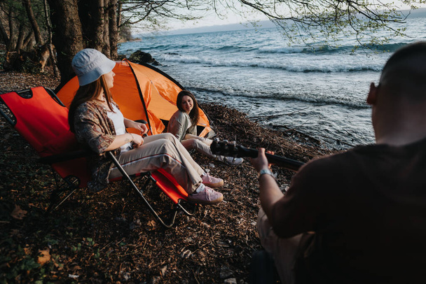Group of friends gathered by a lake, relaxing and having fun at a campsite with a tent and chairs - Photo, Image