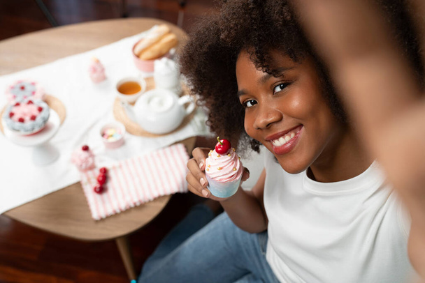 Kaunis nuori afrikkalainen bloggaaja esittelee pala cupcake käsite erityistä ruokaa selfie älypuhelimella. Sisältö luominen sosiaalisen median suosikki makeisia leipomo ruokalaji. Maustevalmistaja. - Valokuva, kuva