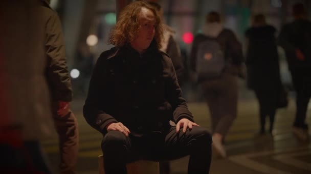 Mężczyzna z długimi włosami w odzieży sportowej siedzi na ławce w nocy na ulicy miasta, podczas gdy grupa ludzi w formalnym stroju przechodzi obok. Inna osoba przechodzi przez drogę w ciemność. - Materiał filmowy, wideo