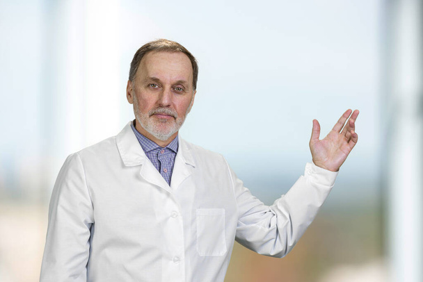 Portrait of senior doctor pointing up showing on copy space. Indoor blurred background. - Photo, Image