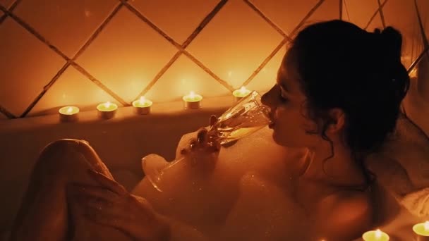 νεαρή κοπέλα με ένα ποτήρι σαμπάνια χαλαρώνει και πίνει ένα αλκοολούχο ποτό στο μπάνιο με αφρό σε ένα ρομαντικό περιβάλλον με ένα αμυδρό φως από το φως των κεριών. Αργή κίνηση - Πλάνα, βίντεο