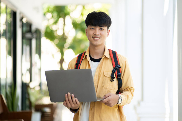 Un estudiante masculino con una sonrisa agradable está parado en el pasillo de la escuela, sosteniendo con confianza su computadora portátil, listo para la clase. - Foto, imagen