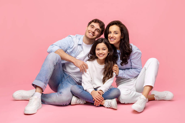 In voller Länge Aufnahme der schönen europäischen Familie sitzt auf rosa Hintergrund, umarmt und genießt die Zeit miteinander zu verbringen - Foto, Bild