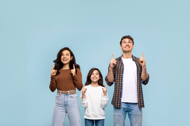 Joyful parents and girl looking and pointing up at free space, standing over blue background, showing place for ad above head, banner - Photo, Image
