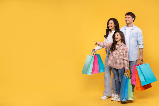 Oferta de compras. Familia europea con hija llevando bolsas de compras y apuntando al espacio libre sobre fondo amarillo, disfrutando de ventas estacionales - Foto, imagen