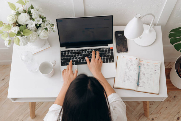 Top kilátás barna nő gépelés szöveget laptop fehér irodai asztal asztali lámpa, notebook, üveg víz és virágok vázában. A szabadúszó, távmunka fogalma otthon, online tanulás - Fotó, kép