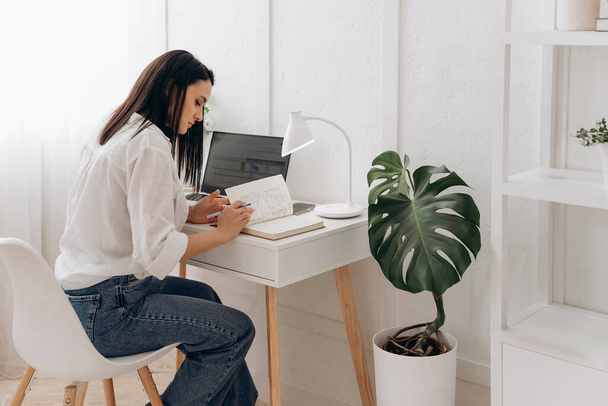 Młoda brunetka freelancer kobieta siedzi przy biurku za pomocą notatek pisania laptopa podczas nauki online, oglądania webinarium, patrząc na PC screen learning web classes lub zdalnej pracy z domu. Widok boczny. - Zdjęcie, obraz