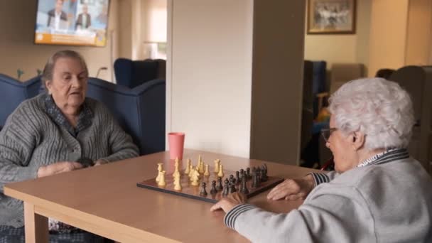 Vanhemmat naiset puhuvat pelatessaan shakkia yhdessä pöydässä kuin viettää vapaa-aikaa ruokalassa hoitokodissa - Materiaali, video