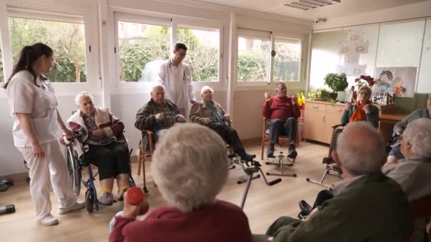 Young male doctor talking to aged man working out using bike pedals and lifting dumbbells during rehabilitation session in retirement home - Footage, Video