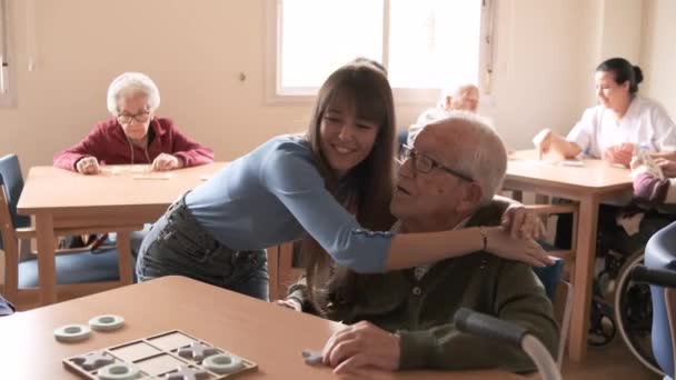 Zeitlupe der jungen glücklichen Enkelin, die einen alten Mann in Brille umarmt und küsst, der im Altersheim am Tisch sitzt - Filmmaterial, Video