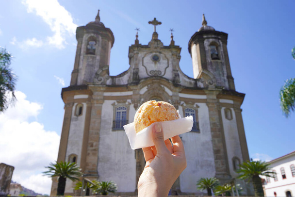 Pao de queijo (pan de queso brasileño) con la iglesia de Nuestra Señora del Monte Carmelo en Ouro Preto, Minas Gerais, Brasil, la ciudad es Patrimonio de la Humanidad por la UNESCO - Foto, Imagen