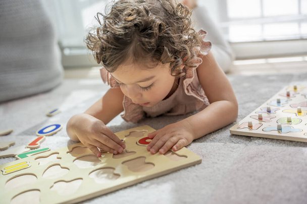 Közelkép egy kislányról, aki egy játékszőnyegen fekszik, és egy fa ábécé betűs puzzle-lel játszik. Montessori módszertan és anyagok. oktatás, fejlesztés és tanulás a gyermekkorban. - Fotó, kép