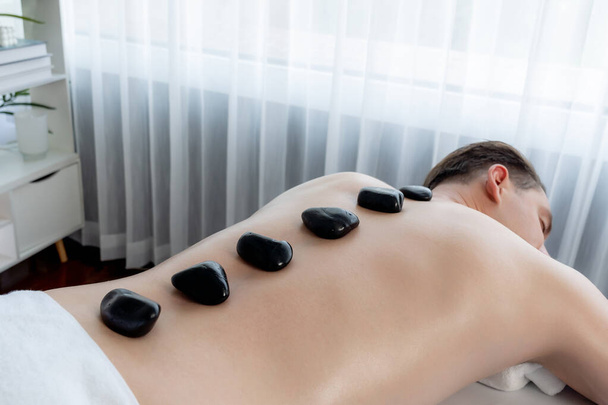 Hot stone massage at spa salon in luxury resort with day light serenity ambient, blissful man customer enjoying spa basalt stone massage glide over body with soothing warmth. Quiescent - Photo, Image