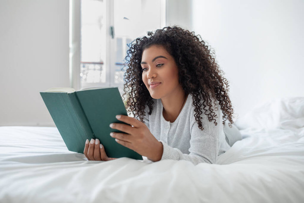 La mujer hispana está sentada en una cama con un libro en las manos, absorta en la lectura. La habitación es acogedora y bien iluminada, creando un ambiente de lectura cómodo. - Foto, Imagen