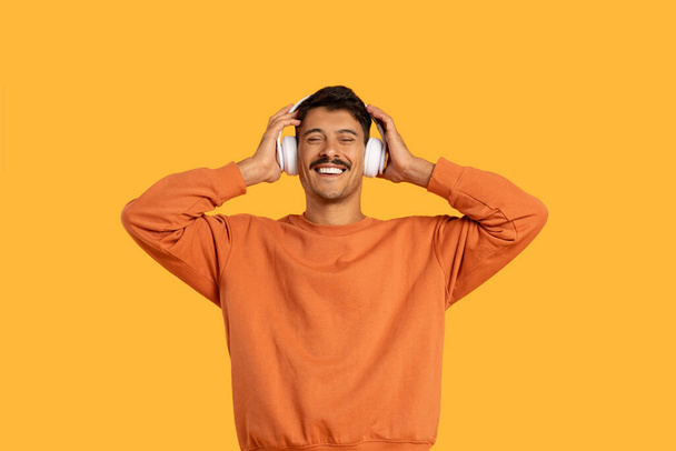 Чоловік у помаранчевій сорочці слухає навушники, зосереджуючись уважно на відтворенні музики. Його вираз роздумує, коли він насолоджується звуком. - Фото, зображення