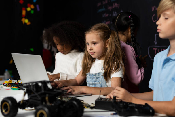 Έξυπνο κορίτσι με λευκή σαλιάρα που μαθαίνει πώς να κωδικοποιεί την τεχνολογία ρομποτικής χρησιμοποιώντας φορητό υπολογιστή στην τάξη STEM. Schoolboy σε μπλε πουκάμισο προσπαθούν να εκπαιδεύσουν μητρική πλακέτα, ενώ έξυπνες μαθήτριες ανάγνωση κώδικα. Εκπομπή. - Φωτογραφία, εικόνα