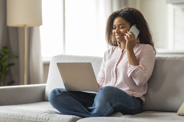 Questa immagine raffigura una giovane donna di colore su un divano, profondamente impegnata in una conversazione telefonica mentre contemporaneamente lavora sul suo computer portatile, mostrando un ambiente dinamico di lavoro da casa - Foto, immagini