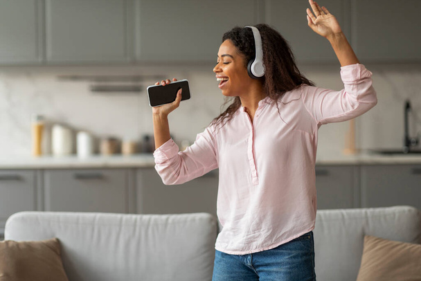 Happy young black woman with headphones singing and dancing at home, using smartphone as microphone - Photo, Image