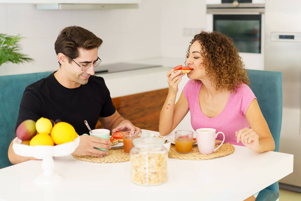 Aikuinen pari rento vaatteet katselee alas, kun istuu ruokapöydässä kupit mehu lasit placemat hedelmiä kulhoon ja syö terveellistä aamiaista päivänvalossa keittiössä - Valokuva, kuva