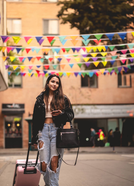 Stylish travel and urban exploration: vibrant young latina brunette with fashion look and pink suitcase exploring city downtown looking map on phone - Photo, Image