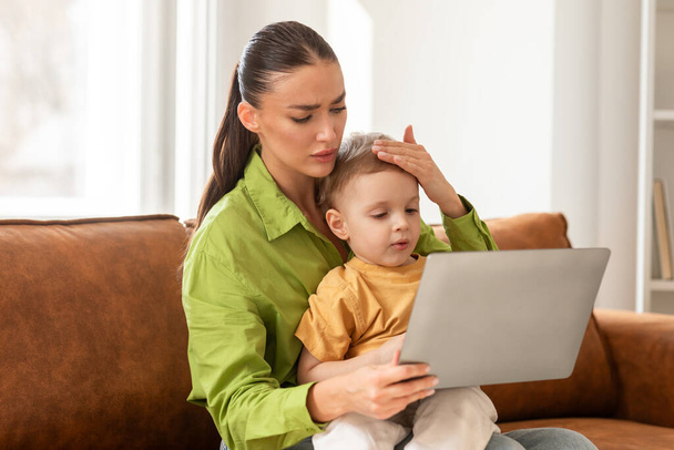 A concerned young woman, dressed in a casual green shirt, is seated on a comfortable sofa as she gently places her hand on her child forehead, seemingly checking for a fever - Photo, Image
